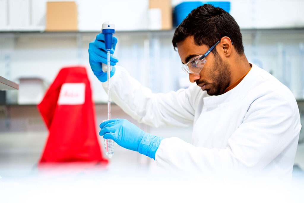 Lab technician wearing safety glasses and blue gloves using a pipette to extract a clear liquid from a beaker.