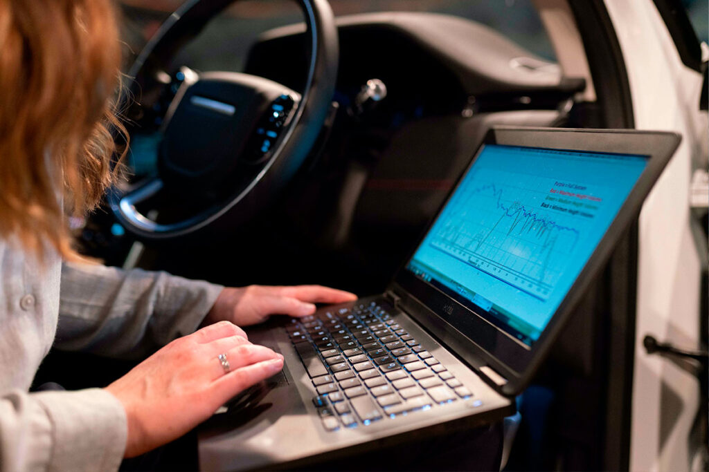 Person with long hair sitting in the driver seat of a car typing on a computer. The computer screen shows a line graph.